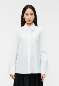 Λευκό πουκάμισο με κουμπιά, γιακά, μακριά μανίκια και φαρδιές μανσέτες· λεία υφή, κλασικός σχεδιασμός, φορεμένο με μαύρη φούστα.