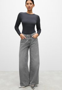 Langsleeve donkergrijze bodysuit gecombineerd met hoge taille, wijdvallende vervaagde grijze jeans. Glanzende puntige zilveren hakken. Eentonige achtergrond.