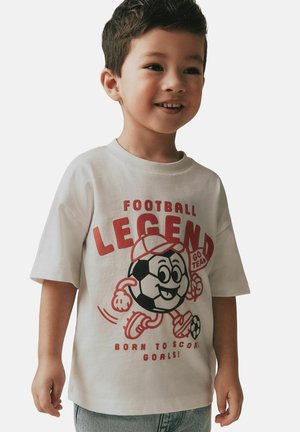 Naeratav noor poiss, kes kannab valget t-särki, millel on joonismängupalli tegelane ja punane tekst "Jalgpallilegendi sündinud väravate löömiseks".