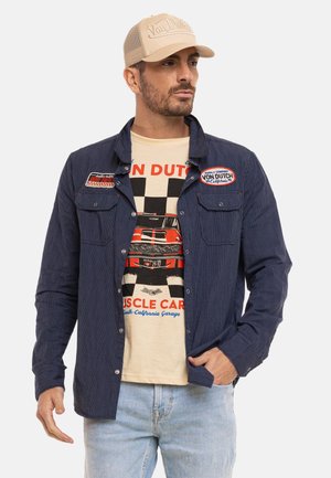 Homme portant une casquette beige Von Dutch, une chemise boutonnée bleu marine et un T-shirt beige avec un graphique de muscle car rouge et un drapeau à damier, associé à un jean bleu clair.