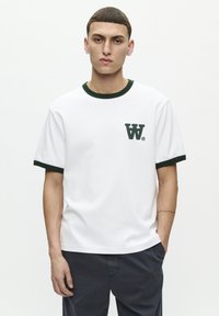 White cotton t-shirt with green trim around the collar and sleeves, featuring a small green logo on the left chest. Short sleeves and relaxed fit.