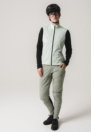 Person trägt hellgrüne Radweste über schwarzen langärmligen Shirt, graue Hose, schwarzen Helm, Brille und schwarze Schuhe und steht vor einem einfarbigen Hintergrund.