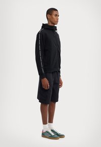 Sudadera con cremallera negra y rayas blancas en los laterales, combinada con pantalones cortos negros y zapatillas verdes con detalles rosas. El estilo es casual y deportivo.