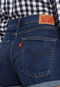 Jean en denim bleu foncé avec une étiquette logo Levi's rouge, des poches arrière hautes avec une couture distinctive, et un patch de marque en cuir marron.