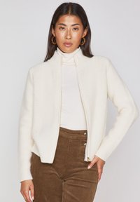 Cardigan zip-up color crema in maglia texture, con colletto alto e polsini a coste, abbinato a pantaloni in velluto a coste marroni.