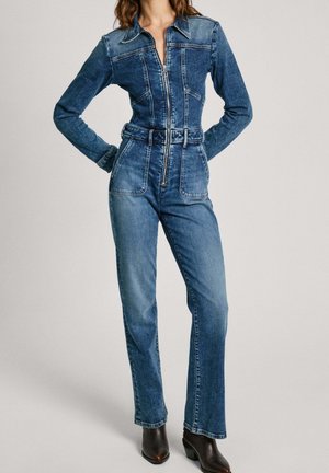 Femme portant une combinaison en denim cintrée à manches longues avec fermeture éclair à l'avant, taille ceinturée et pantalon droit, associée à des bottines foncées.