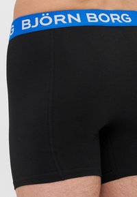 Black boxer shorts featuring a blue elastic waistband with "BJÖRN BORG" in white text, highlighting a smooth texture and fitted design.