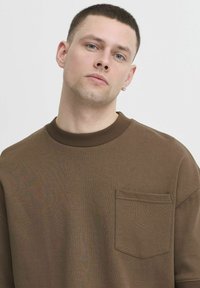Brun, ribbet genser med rund hals og korte ermer, med en lomme på venstre bryst. Glatt tekstur med en tettsittende krage.