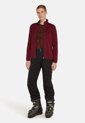 Maroon zip-up fleece jacket with high collar, paired with a black patterned top and black ski pants. Black ski boots with green accents.