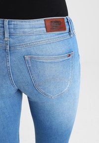 Vaaleansiniset farkut, joissa on keskimäinen vyötärö, ruskea nahkapaikka, jossa lukee "TOMMY JEANS", ja pieni logo takataskussa.