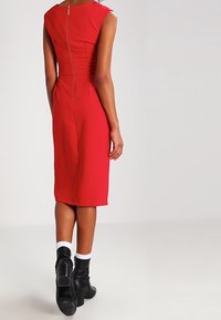 Robe rouge ajustée à manches courtes, fermeture éclair au dos. Portée avec des bottines noires et des chaussettes blanches. Texture lisse, coupe au niveau des genoux.