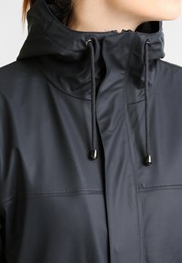 Czarna wodoodporna kurtka z wysokim kołnierzem, regulowanymi sznurkami, gładką fakturą i minimalistycznym wzornictwem. Z przodu zamek błyskawiczny, bez widocznych wzorów.