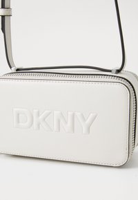 Bílá crossbody taška DKNY s embosovaným logem, obdélníkový tvar, hladká textura, černý zip a kontrastní černý popruh.