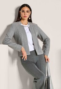 Vrouw in een grijs gestructureerd blazer, wit top, grijze broek, grote heldere bril, paarse oorbellen met parels, die een zilveren handtas vasthoudt.