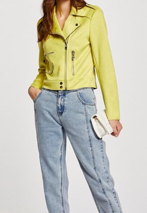 Femme portant une veste courte jaune vif avec des détails zippés, un jean taille haute bleu clair, et tenant une petite pochette blanche.