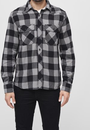 Homme portant une chemise à carreaux noire et grise à boutons avec deux poches poitrine et un pantalon noir, debout devant un fond uni.