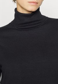 Crna turtleneck džemper u rebrastom pletenju. Ima visoki ovratnik i prilagođeni dizajn, koji ističe glatku teksturu kroz cijeli komad.