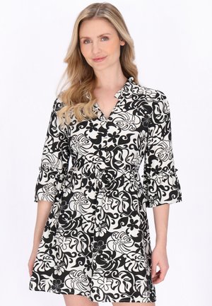 Kvinde med langt blondt hår iført en knælængde kjole i sort og hvidt med blomstermønster og abstrakte detaljer samt trekvartlange ærmer.