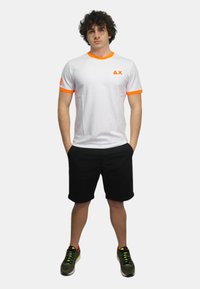 T-shirt bianca a maniche corte con dettagli arancioni sul colletto e sulle maniche, caratterizzata da un logo sul petto. Indossata con pantaloni corti neri e scarpe sportive.