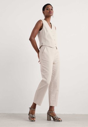 Beige ärmellose Weste mit Knöpfen, kombiniert mit hellbeigen, maßgeschneiderten Hosen. Das Modell trägt metallische High-Heels. Glatte Textur.