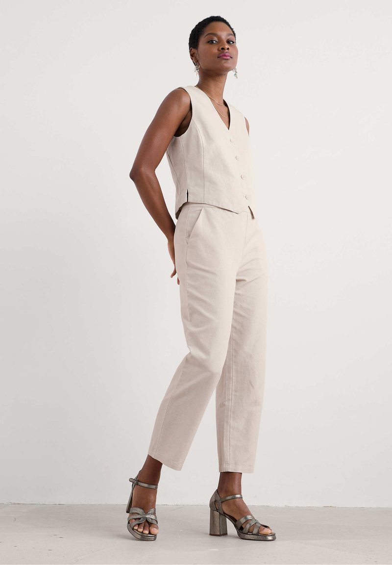 Beige ärmellose Weste mit Knöpfen, kombiniert mit hellbeigen, maßgeschneiderten Hosen. Das Modell trägt metallische High-Heels. Glatte Textur.