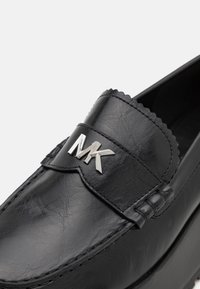Mocassin en cuir noir avec logo "MK" argenté sur la sangle, avec des coutures détaillées et un design à bords festonnés.