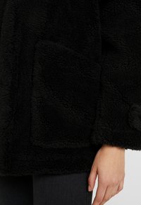 Czarna, włochata odzież wierzchnia o teksturowanej powierzchni, z przednią kieszenią naszywaną. Zauważ luźny krój i miękki, ciepły materiał.