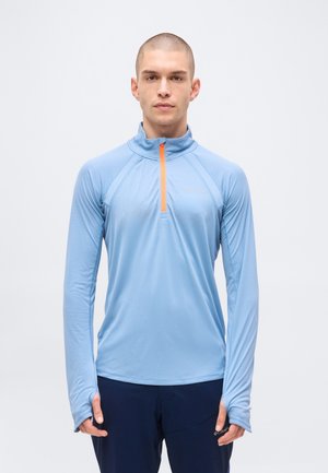Homme aux cheveux courts portant un haut de sport bleu clair à manches longues avec une fermeture éclair orange et un pantalon foncé, debout devant un fond uni.