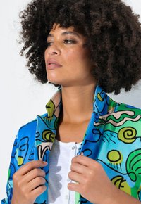 Veste à motifs bleus et verts avec de grands designs abstraits et un col haut, portée sur un haut blanc, mettant en valeur une texture de tissu lisse.