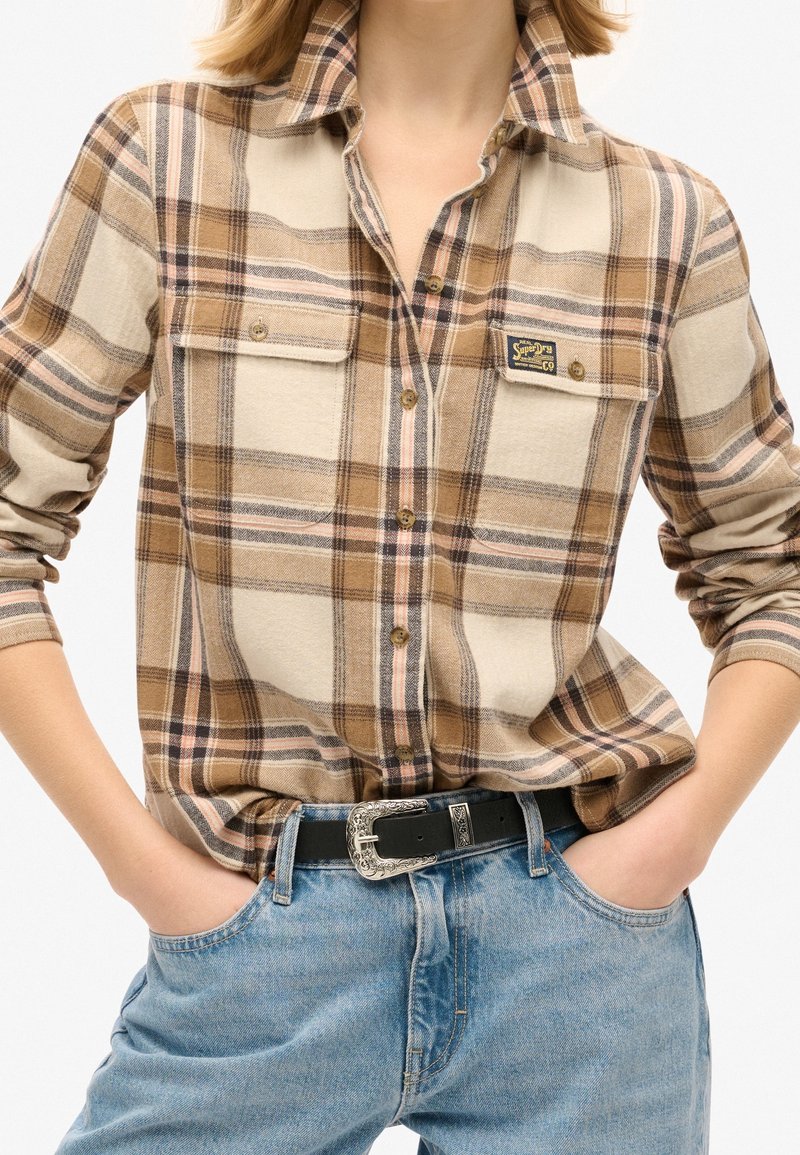 Personne portant une chemise à carreaux beige et marron avec des manches retroussées, rentrée dans un jean bleu clair avec une ceinture noire.