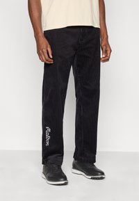 Pantalon en velours côtelé noir avec des côtes verticales, arborant un logo blanc brodé sur la jambe gauche. Porté avec une chemise courte beige et des chaussures noires.