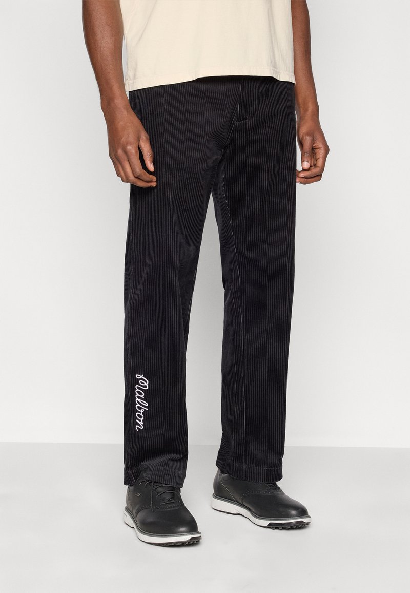 Pantalon en velours côtelé noir avec des côtes verticales, arborant un logo blanc brodé sur la jambe gauche. Porté avec une chemise courte beige et des chaussures noires.