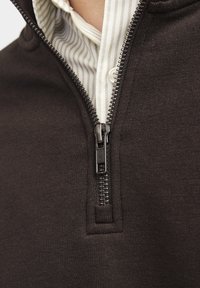 Sudadera con cremallera en tejido de punto marrón oscuro con un acabado texturizado, que presenta una cremallera negra y una camiseta interior a rayas en tonos claros.