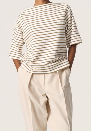 Persoon draagt een loszittend wit en beige gestreept T-shirt met korte mouwen, ingestopt in een hooggetailleerde, crèmekleurige geplooide broek, handen in de zakken.