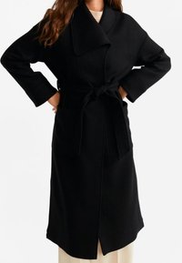 Černý vlněný kabát s velkým límcem, širokými rukávy, páskem v pase a předním rozparkem, s hladkou texturou a minimalistickým designem.