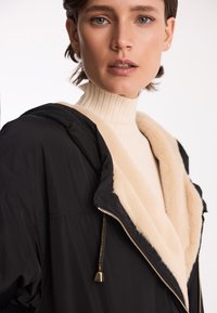 Czarna kurtka z kapturem z beżowym kołnierzem z futra syntetycznego. Posiada zamek błyskawiczny, sznurek oraz miękką teksturę. Nosi się ją na prążkowanym kremowym golfie.