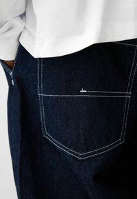 Tamno traperice s bijelim šavovima na stražnjem džepu. Tkanina pokazuje glatku teksturu s strukturiranim dizajnom.