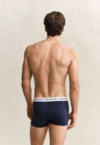 Homme debout de dos, portant un boxer bleu marine avec une ceinture blanche portant la marque GANT sur un fond uni.