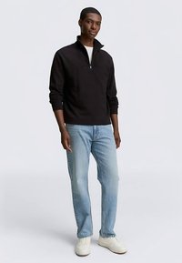 Fekete negyedcipzáras pulóver hosszú ujjakkal, világoskék egyenes szárú farmernadrággal és fehér tornacipővel párosítva. Sima szövettextúra.