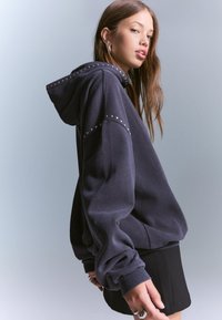 Hanorac gri închis cu o croială lejeră, decorat cu studs metalici pe umeri și cu o glugă mare. Material neted, cu manșete ribbate.