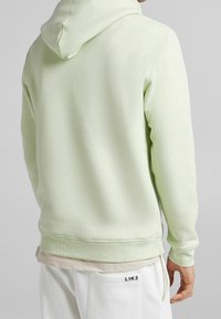 Felpa verde chiaro con una texture morbida, dotata di cappuccio con cordino e polsini a coste. Indossata sopra un indumento beige e abbinata a pantaloni bianchi.