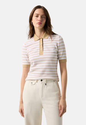 Kobieta o brązowych włosach nosi beżowo-białą, krótki rękaw koszulkę polo w paski oraz jasnobeżowe spodnie z wysokim stanem, stojąc na białym tle.
