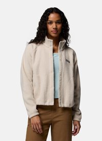 Woman with curly hair wearing a beige fleece jacket over a light blue top and brown pants, standing against a plain background.