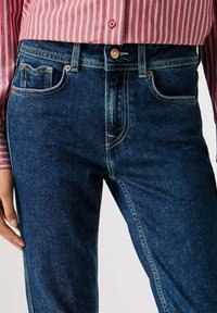 Tumši zili džinsi ar taisnu griezumu, jostas cilpām un redzamām šuvju detaļām. Ir priekšējā kabata ar klasisku dizainu.