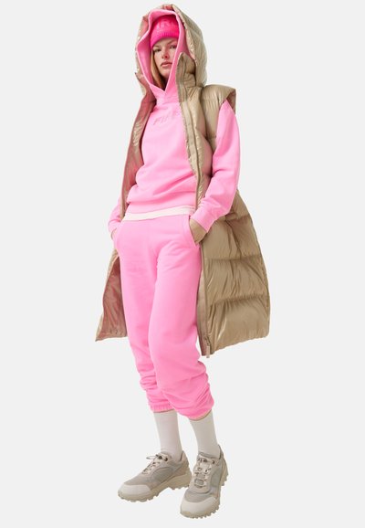 Wattierte Weste in metallic Beige, kombiniert mit einem pinken Hoodie und Trainingshose. Dazu gehören klobige graue Sneaker und weiße Socken.