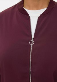 Burgunderfarbene Zip-Jacke aus glattem Stoff, mit Rundhalsausschnitt, silbernem Reißverschluss und einer weißen inneren Lage, die am Kragen sichtbar ist.