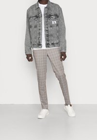 En person står i en grå denimjacka över en vit skjorta, tillsammans med beiga rutiga byxor och vita sneakers mot en enhetlig bakgrund.