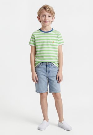 Αγόρι που στέκεται, φοράει πράσινη και λευκή ριγέ μπλούζα, ανοιχτόχρωμα τζιν σορτς και λευκά παπούτσια slip-on, με φόντο απλό λευκό.