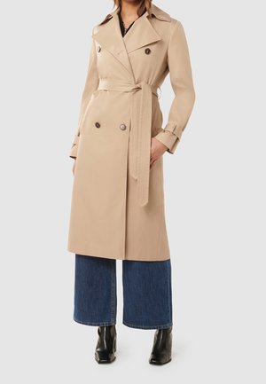 Femme portant un trench-coat beige croisé avec ceinture, un jean large bleu et des bottines noires, debout devant un fond clair.
