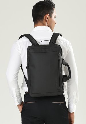 Uomo che indossa una camicia bianca e pantaloni neri, con uno zaino rettangolare nero dotato di tasche con cerniera laterale e frontale, visto di spalle.